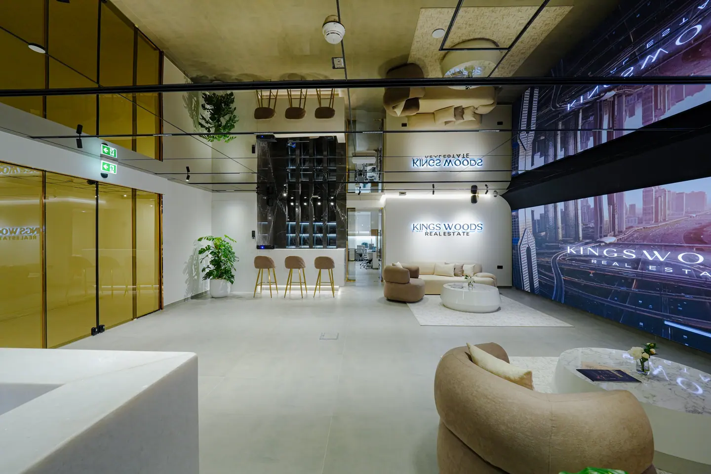 Open lounge and waiting area with gold-framed glazing, bar stools, planting and a branded video wall — Kings Woods Real Estate office fit-out by Urban Crown, Dubai